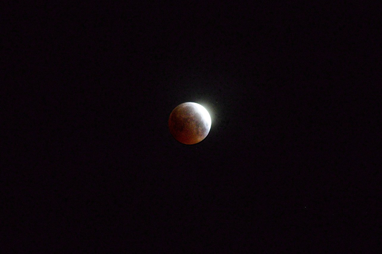 A Moon captured during a lunar eclipse against a dark night sky. The left portion of the Moon displays a reddish-brown coloration typical of a blood moon, while the right edge remains brightly illuminated, creating a striking crescent of light. A subtle glow surrounds the Moon, enhancing its ethereal appearance in the otherwise black expanse.